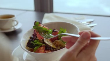 Kadın, deniz manzaralı pencerenin yanında oturmuş domatesli fırında patlıcan yiyor.