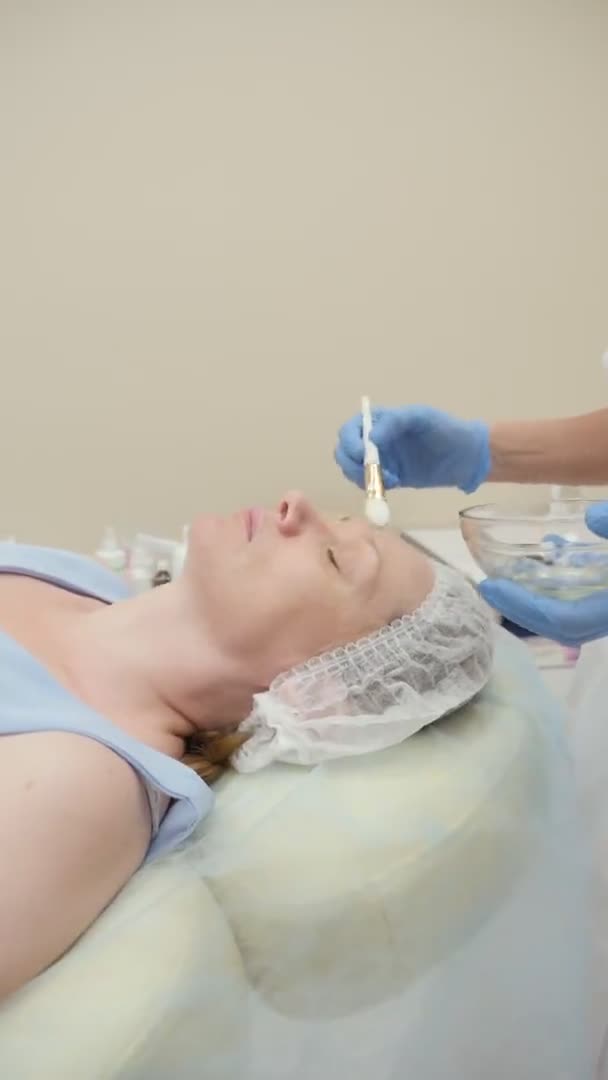 Dikey video. Yakın plan. Güzellik uzmanı bir kadının yüzüne fırçayla maske takar. — Stok video