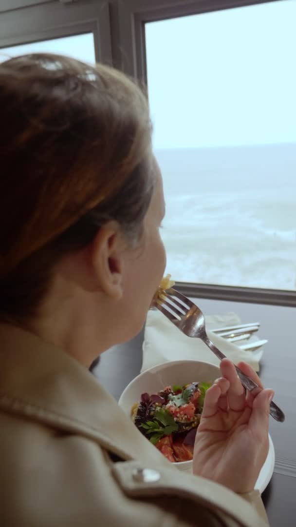 Vertikální video. žena jíst pečené lilku s rajčaty, zatímco sedí u stolu u okna s výhledem na moře — Stock video