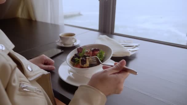 3.女人坐在窗边俯瞰大海的桌子旁边，一边吃着烤着的西红柿茄子 — 图库视频影像