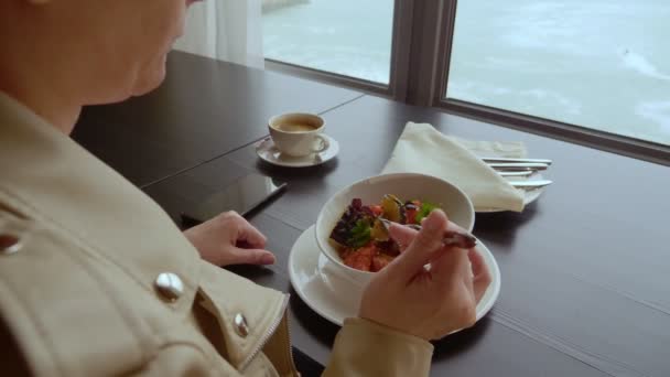 Mujer comiendo berenjena al horno con tomates mientras está sentada en la mesa junto a la ventana con vistas al mar — Vídeo de stock