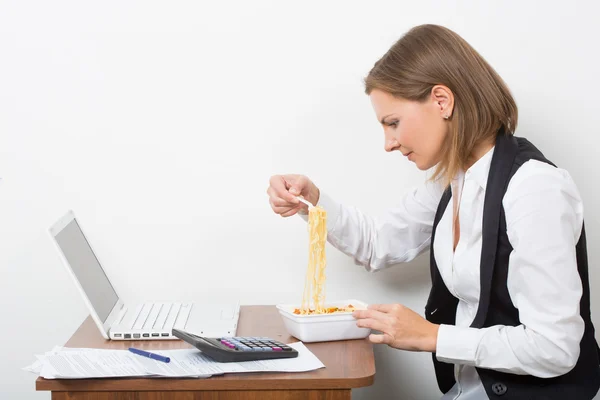 Tjej äter en pasta, arbetar bakom den bärbara datorn. — Stockfoto