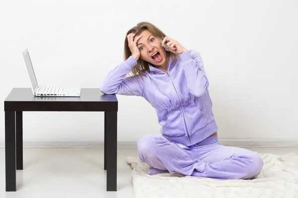 Chica sorprendida hablando por teléfono mientras estaba sentada en casa. junto a un ordenador portátil —  Fotos de Stock