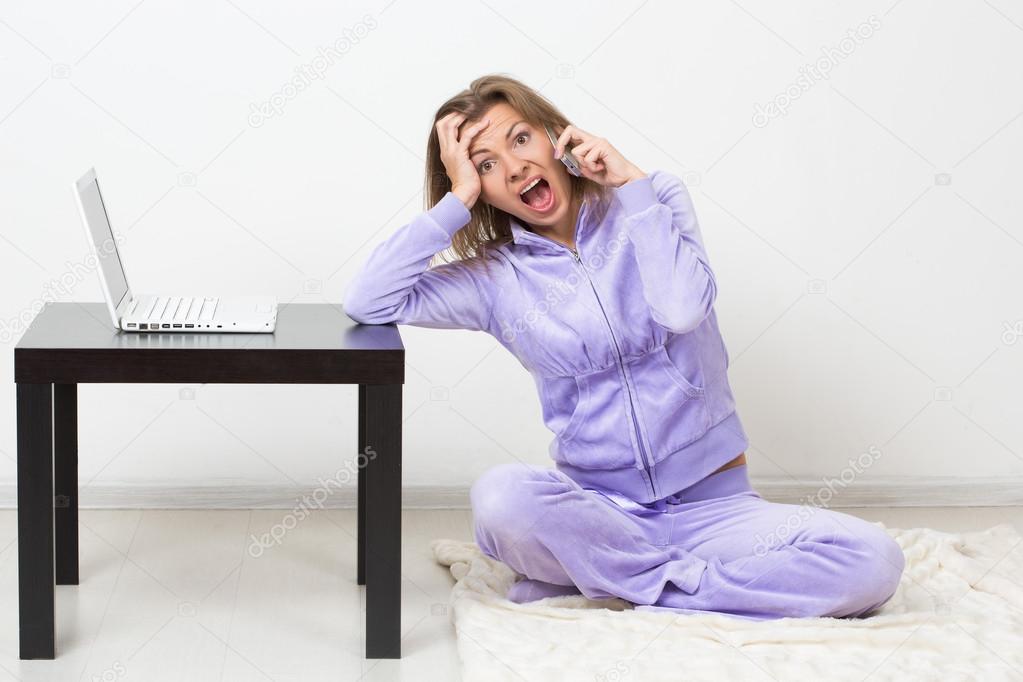 urprised girl talking on the phone while sitting at home. next to a laptop