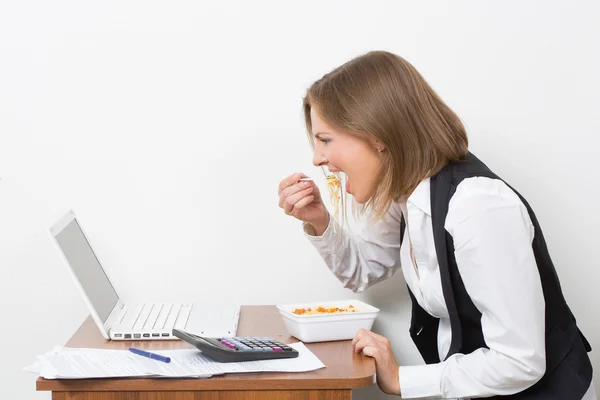 Κορίτσι τρώει ένα ζυμαρικά, εργάζονται πίσω από το φορητό υπολογιστή. — Φωτογραφία Αρχείου