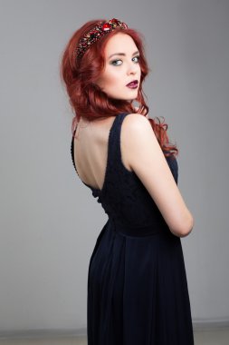 Beautiful red-haired fashion model posing in evening dress and in the diadem over dark background. female gestures of seduction. body language. view from behind the shoulder