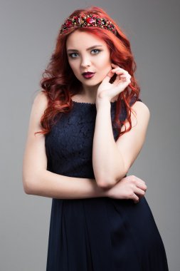 Beautiful red-haired fashion model posing in evening dress and in the diadem over dark background. female gestures of seduction. body language. showing wrist.