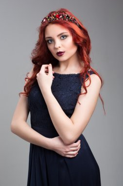 Beautiful red-haired fashion model posing in evening dress and in the diadem over dark background. female gestures of seduction. body language. showing wrist.