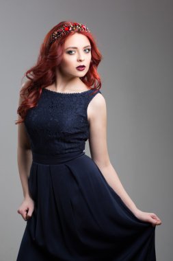 Beautiful red-haired fashion model posing in evening dress and in the diadem over dark background. Wavy Red Hair. Fashion Girl Portrait.