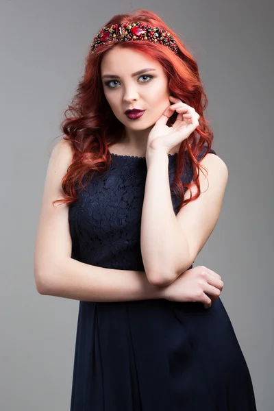 Beautiful red-haired fashion model posing in evening dress and in the diadem over dark background. female gestures of seduction. body language. showing wrist. — Stock fotografie