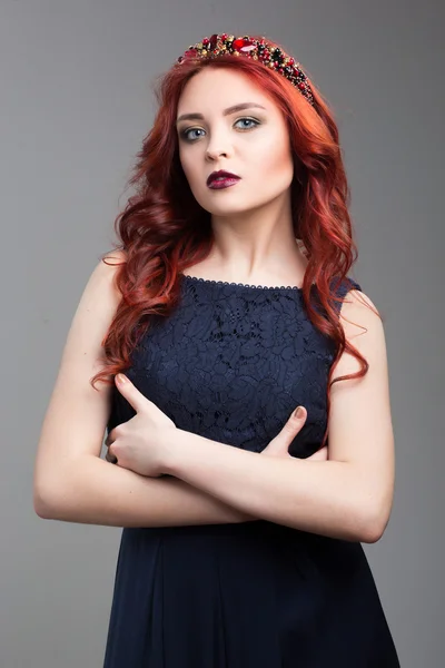 Beautiful red-haired fashion model posing in evening dress and in the diadem over dark background. body language. thumbs demonstration. gesture of superiority. crossed arms — Stock fotografie