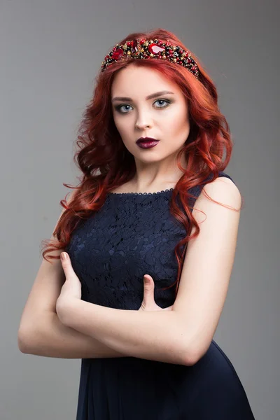 Beautiful red-haired fashion model posing in evening dress and in the diadem over dark background. body language. thumbs demonstration. gesture of superiority. crossed arms — Stok fotoğraf