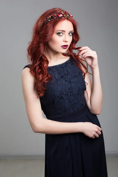Beautiful red-haired fashion model posing in evening dress and in the diadem over dark background. female gestures of seduction. body language. touching her hair. Wavy Red Hair. Fashion Girl Portrait. — Stockfoto