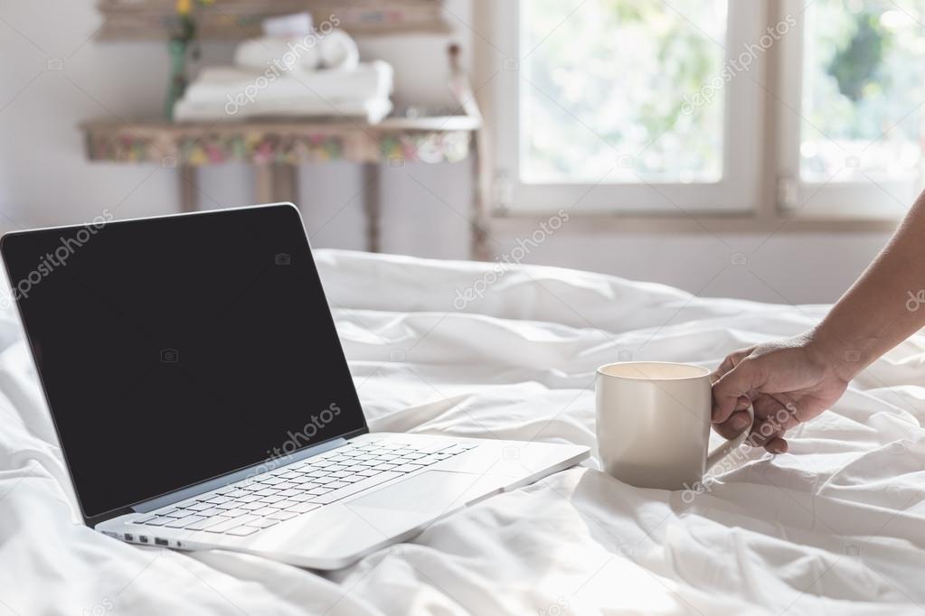 Hand holding coffee cup and laptop on the bed in morning time, f