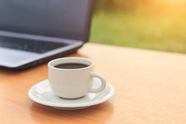 Bliska filiżanki kawy i laptopa na stole w godzinach porannych — Zdjęcie stockowe