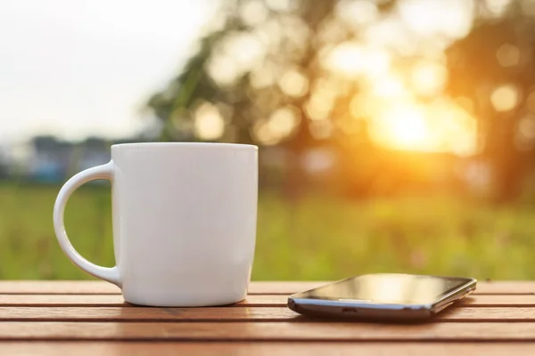 Taza de café y teléfono inteligente en la mesa en la hora del atardecer — Foto de Stock