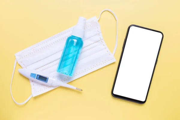 Phone mockup, Alcohol spray, Digital thermometer and Face mask on yellow background.