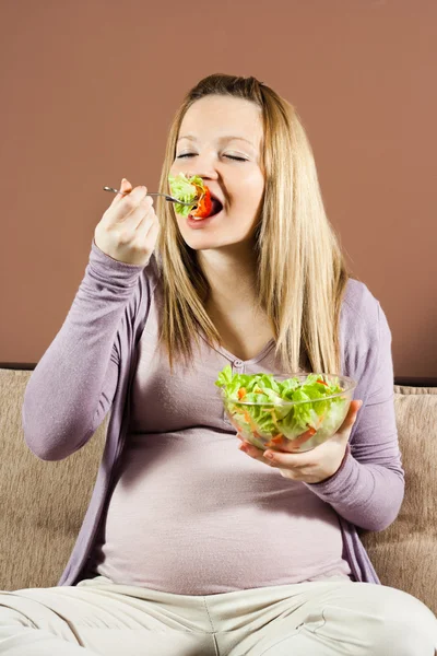 Mujer embarazada sentada en el sofá comiendo ensalada —  Fotos de Stock