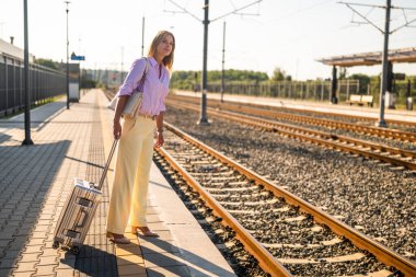 Tren istasyonunda kendine güveni tam bir iş kadını, iş gezisi sırasında bir tren beklerken başka yere bakıyor ve bavullarını tutuyor, seyahat, başarı ve profesyonel hareketliliği simgeliyor..