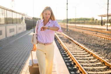 Tren istasyonunda yürürken kol saatine bakan bavullu, kendine güvenen ve zarif bir iş kadını. Modern seyahat, zaman yönetimi ve şehir hayatının sembolü.