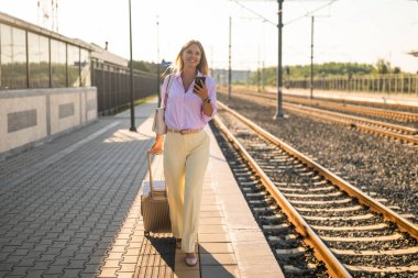 Bavullu güzel bir iş kadını akıllı telefonunu kullanırken demiryolu platformu boyunca yürüyor. Kendine güveni tam, şık ve iş gezisi sırasında hareket halinde..