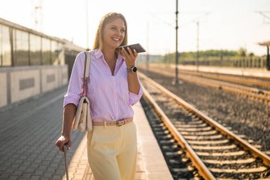 Bavuluyla ayakta duran ve tren istasyonunda akıllı telefon kullanan zarif bir iş kadını. Günlük seyahat, verimlilik ve modern yaşam tarzı kavramı.