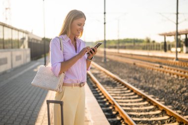 Güzel ve zarif bir iş kadını, tren istasyonunda elinde bir bavulla trenin gelmesini beklerken akıllı telefon kullanıyor.