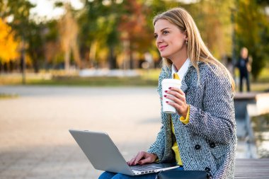 Şık sonbahar montlu modern iş kadını şehirde bir bankta otururken dizüstü bilgisayar kullanıyor ve kahve içiyor.. 