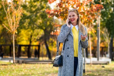 Modern ceketli mutlu iş kadını portresi. Açık havada akıllı telefondan konuşuyor. Etrafı renkli sonbahar yapraklarıyla çevrili..