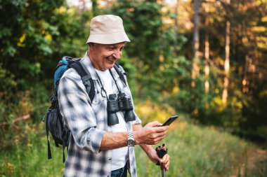 Sırt çantası ve dürbünle gülümseyen yaşlı yürüyüşçünün portresi akıllı telefon kullanarak tek başına dağ yürüyüşü yaparken doğayı keşfediyor..