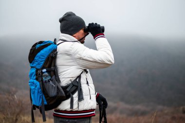 Sırt çantalı, yürüyüş direkleri olan, sisli bir kış gününde doğa yürüyüşü yaparken dürbün kullanan yürüyüşçü, keşfe ve manzaraya dalmış..