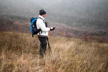 Sırt çantalı, sırıklı ve yürüyüş direkli yürüyüşçü sisli kış günlerinde doğa yürüyüşünün tadını çıkarırken akıllı telefon kullanıyor..