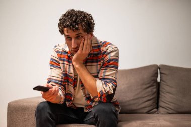 Genç adam evinde kanepede otururken sıkılıyor ve televizyon kanallarını değiştiriyor..