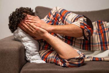 Yorgun ve bunalımda olan genç bir adam ellerini tutuyor ve evinde kanepede uzanıyor..