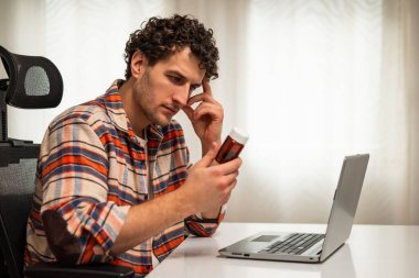 Genç bir adam reçeteli ilaç şişesini tutuyor ve evinde dizüstü bilgisayar kullanıyor. Belirsizlik hissediyor ve ilaç ya da olası yan etkiler için endişeleniyor..