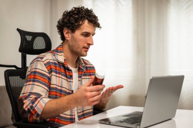 Genç adam dizüstü bilgisayarında doktora ilaç şişesini gösteriyor, evde otururken sağlık durumunu kontrol altına almak için doğru dozun ne olduğunu soruyor. Teletıp kavramı.