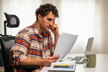 Odaklanmış işadamı rahat evinde çalışırken bilgisayarını kullanıyor ve belgeleri inceliyor..