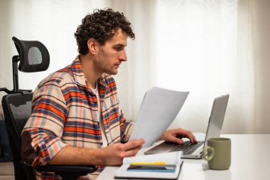 Odaklanmış işadamı rahat evinde çalışırken bilgisayarını kullanıyor ve belgeleri inceliyor..