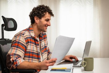 Keyifli genç serbest çalışan. Evinde çalışırken bilgisayarını ve belgelerini kontrol ediyor..