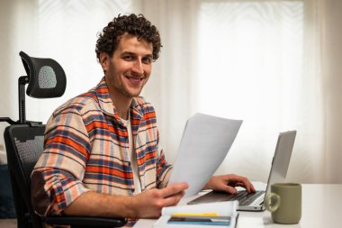 Konforlu evinde çalışırken bilgisayarını kullanan ve belgeleri kontrol eden mutlu genç iş adamının portresi..