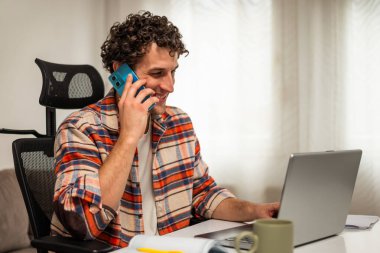 Akıllı telefon ve dizüstü bilgisayar kullanan mutlu genç işadamı rahat evinde çalışmaktan zevk alıyor..