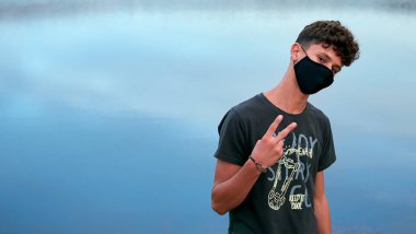 Siyah bir maske takan ve mavi bir gölün önünde poz veren genç beyaz bir adam.