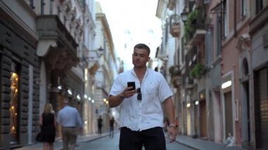 Güzel kalabalık caddede elinde akıllı telefonuyla yürüyen sıradan giysili yakışıklı bir adam. Dışarıdaki muhteşem binaya bakan ve öğleden sonra yürüyüşünden zevk alan genç bir erkek. Kullanılan kişi