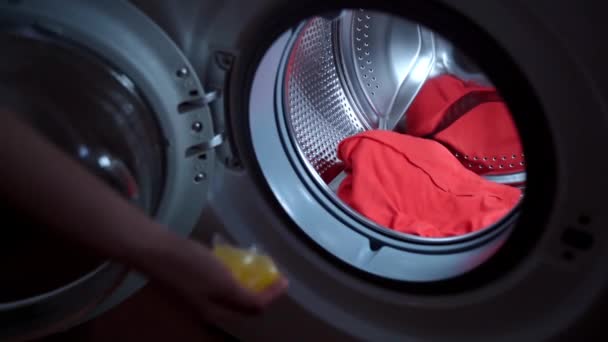 Домогосподарка кидає дві жовті капсули рідкого концентрованого порошку всередину циліндра пральної машини і закриває двері. Жінка готує брудну білизну для прання, жовтий рідкий засіб для прання — стокове відео