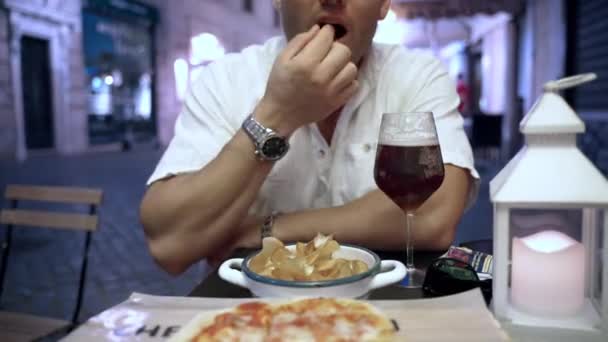 Hungry man sitting outdoors at the table in local pub and eating potato chips, delicious italian pizza and drinking beer. Unhealthy food in modern society, fast food concept — Stock Video