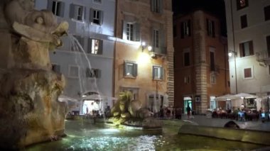 Ağzı açık bir balık heykeli ve şehir merkezindeki antik mermer Roma çeşmesinde akan su. Aydınlatılmış antik çeşme ve geceleri kalabalık cadde, Roma mimari mirası