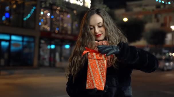 Mujer bastante sonriente con guantes de invierno sosteniendo el regalo de Navidad en las manos y abriéndolo, mujer recibió regalo especial de Navidad de persona querida. Mujer feliz disfrutando de la noche al aire libre en la calle iluminada — Vídeo de stock
