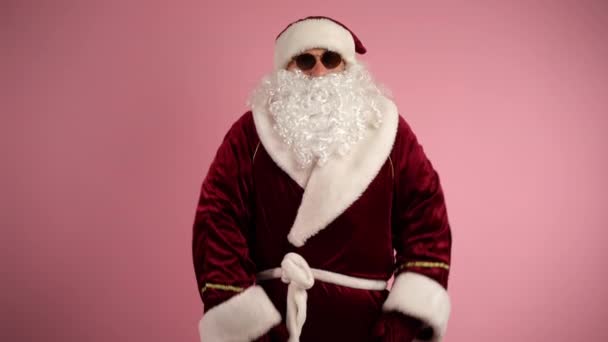 Santa Claus en gafas oscuras y traje tradicional rojo de pie sobre fondo rosa y mirando a la cámara. Santa sorprenderse y lanzar los brazos de ancho, concepto de vacaciones de Navidad y el invierno — Vídeo de stock