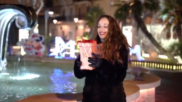 Glad leende kvinna i vinterkläder dansar i parken med jul närvarande i händerna, kvinnliga njuter vintersemester utomhus på natten nära den strömmande fontänen vatten. Uppriktiga känslor av — Stockvideo