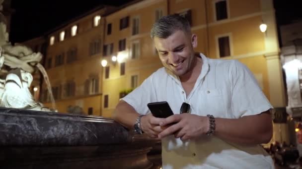 夜晚明亮的罗马街，令人惊奇的建筑外观和微笑的视频内容创造者站在大理石喷泉旁边，并使用手机。积极的男人说话，微笑，聊天 — 图库视频影像
